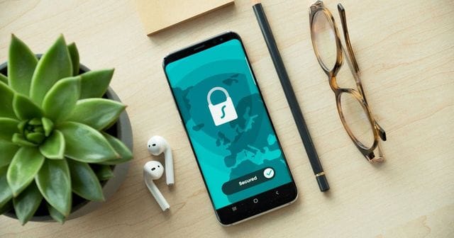 Smartphone con un lucchetto sullo schermo su una scrivania chiara, tra pianta grassa, auricolari e occhiali