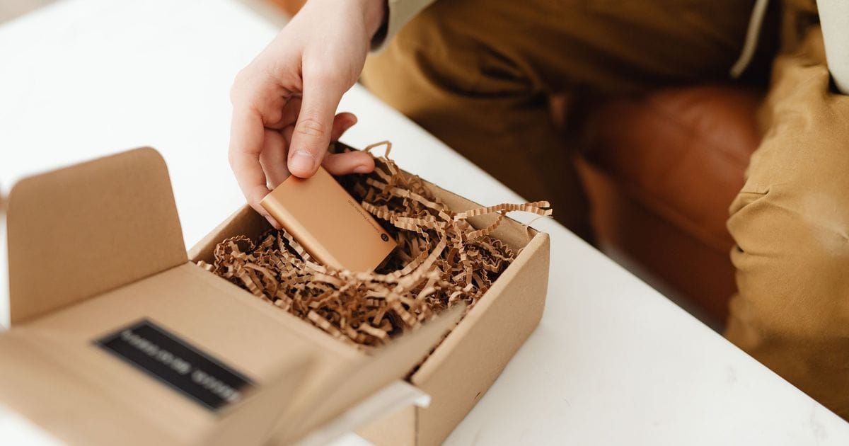 Una mano tira fuori un piccolo oggetto da un pacco di cartone appena aperto, pieno di carta di riempimento