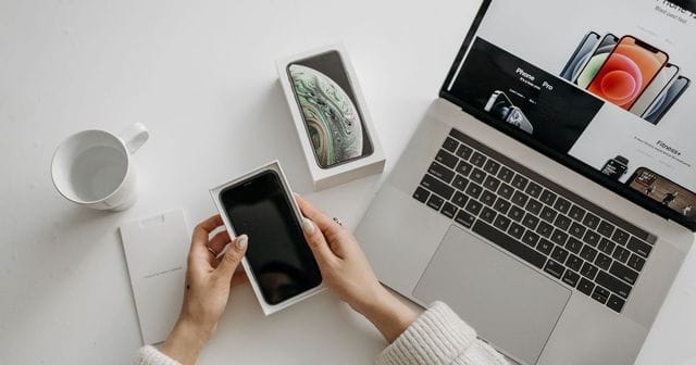 Mani che tolgono uno smartphone dalla confezione accanto a un portatile aperto su una pagina di prodotti