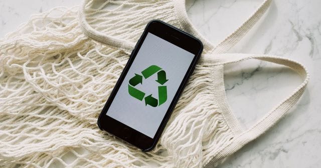 Smartphone con il simbolo del riciclo sullo schermo, appoggiato su una borsa a rete in cotone su un piano di marmo