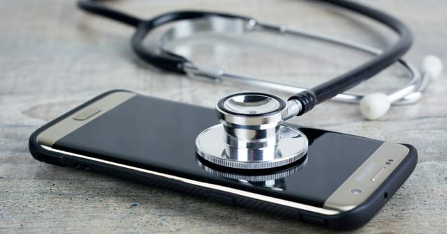 Uno stetoscopio appoggiato sullo schermo di uno smartphone, su un piano di legno grigio