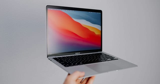 Una mano regge un MacBook Air aperto con lo schermo colorato, su fondo grigio chiaro