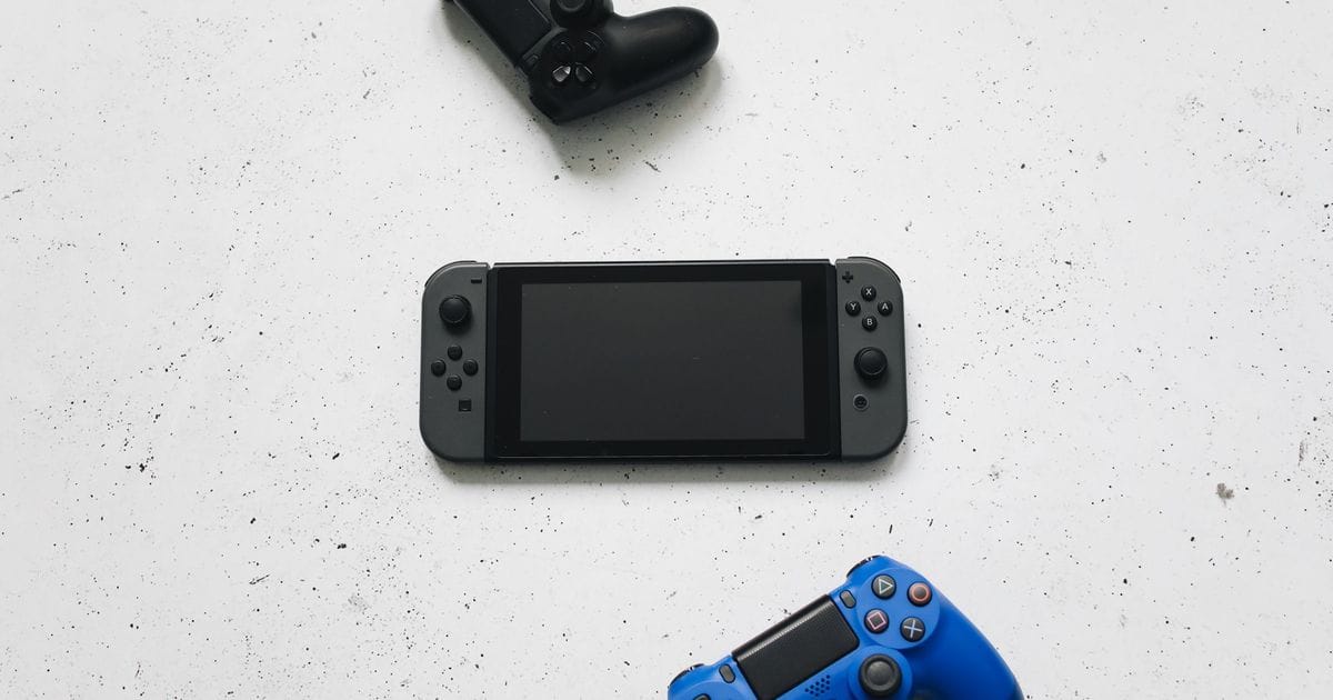 Nintendo Switch vista dall'alto tra due controller PlayStation, uno nero e uno blu, su un piano bianco